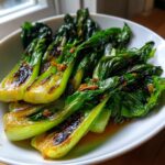 A white bowl filled with glossy, perfectly sauteed bok choy, showing slight charring on the stalks.
