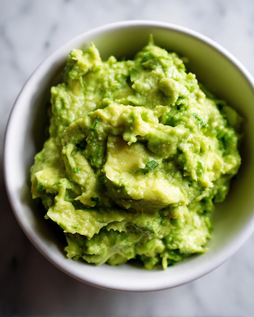 A close-up overhead shot of chunky, bright green authentic guacamole served in a small white bowl.