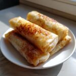 Close-up of four golden, slightly browned baked apple pie rice paper rolls stacked on a white plate.