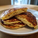Close-up of a stack of golden-brown banana cottage cheese pancakes, one cut in half showing the fluffy interior.