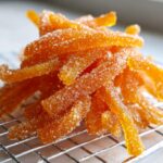 Close-up of bright orange candied orange peel strips generously coated in sparkling sugar, resting on a wire cooling rack.