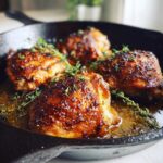 Four golden-brown, crispy skin cast iron skillet chicken thighs garnished with fresh thyme sprigs.