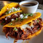 Two crispy, cheesy Beef Birria tacos topped with diced onion and cilantro, served next to a small bowl of dark consommé.