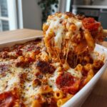 A spoonful of cheesy pizza pasta bake being lifted from the dish, showing long cheese pulls and pepperoni slices.