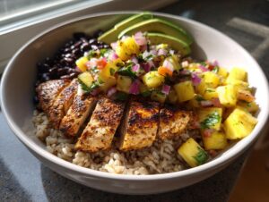 A close-up of a healthy chicken bowl with pineapple salsa, brown rice, black beans, and sliced avocado.