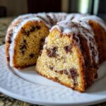 A slice cut from a moist chocolate chip bundt cake, drizzled with white glaze, sitting on a white plate.