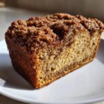 A close-up of a moist slice of coffee cake banana bread featuring a thick, crumbly cinnamon streusel topping.