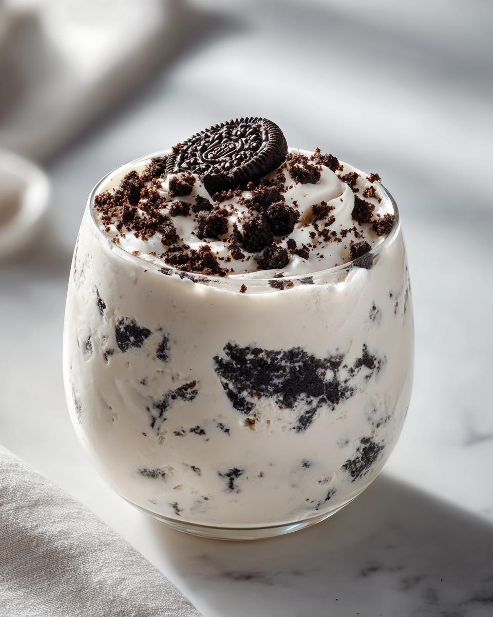 A glass filled with creamy cookies and cream mousse, topped with whipped cream, cookie crumbles, and a whole cookie.