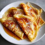 A stack of folded Crepes Suzette drizzled with orange syrup and dusted with powdered sugar on a white plate.