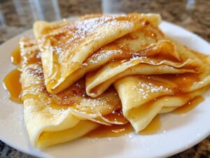 Close-up of folded Crepes Suzette drizzled with orange syrup and dusted with powdered sugar on a white plate.