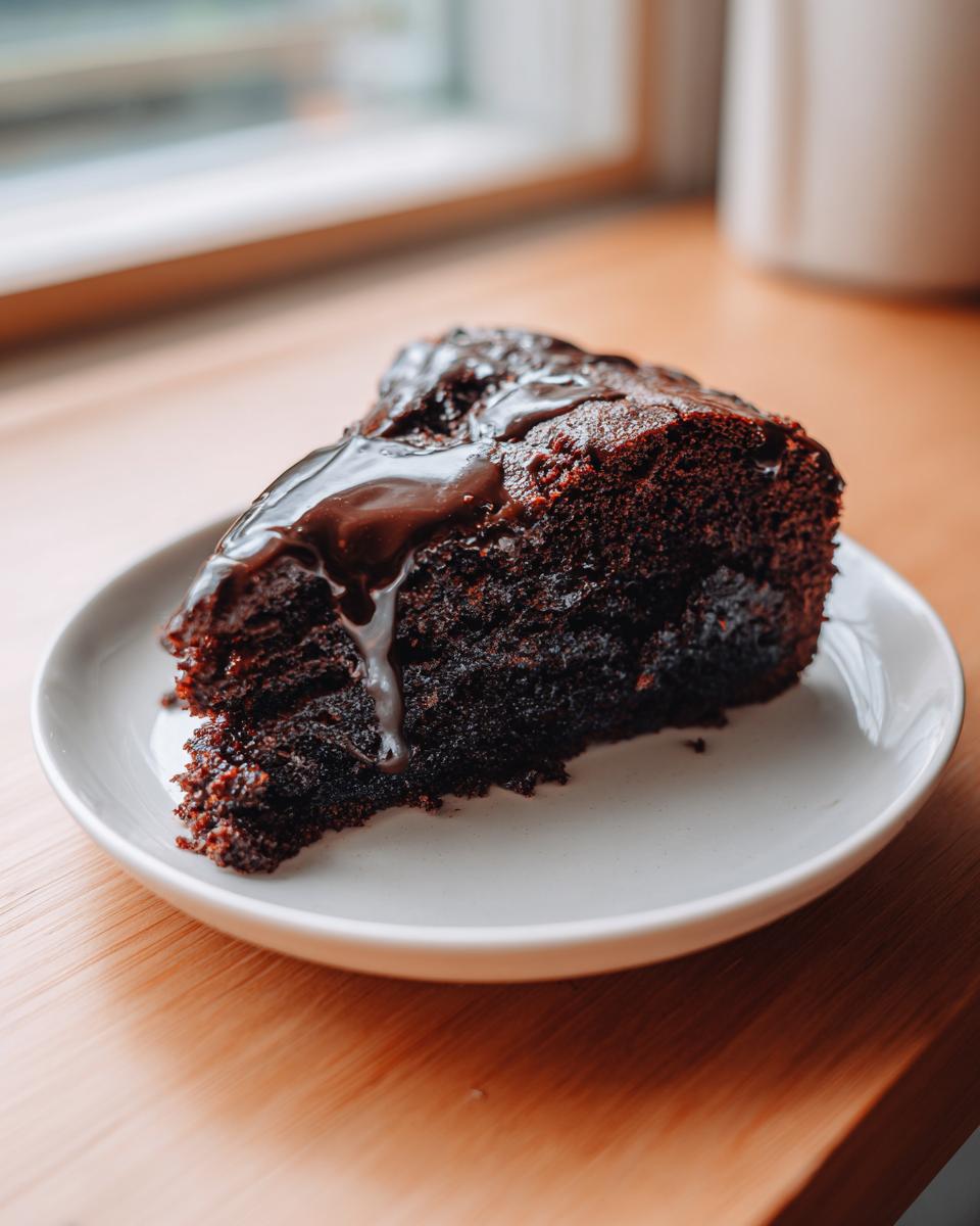A moist slice of dark chocolate dr pepper cake topped with dripping chocolate glaze on a white plate.