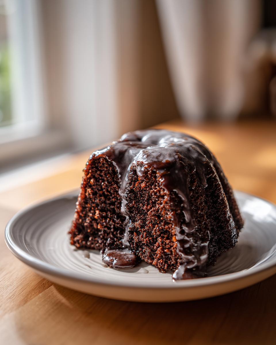 A moist slice of dark chocolate dr pepper cake topped with a thick, glossy chocolate glaze, served on a light plate.