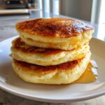 A stack of three thick, fluffy muffin mix pancakes drizzled generously with golden syrup on a white plate.