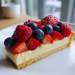 A close-up of a slice of fresh fruit tart topped with strawberries, blueberries, and raspberries, dusted with powdered sugar.