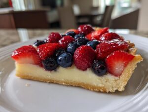 A close-up slice of a fresh fruit tart featuring strawberries and blueberries over a creamy filling.