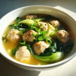 A close-up of a white bowl filled with savory ginger chicken meatball soup, featuring meatballs, bok choy, and green onions.