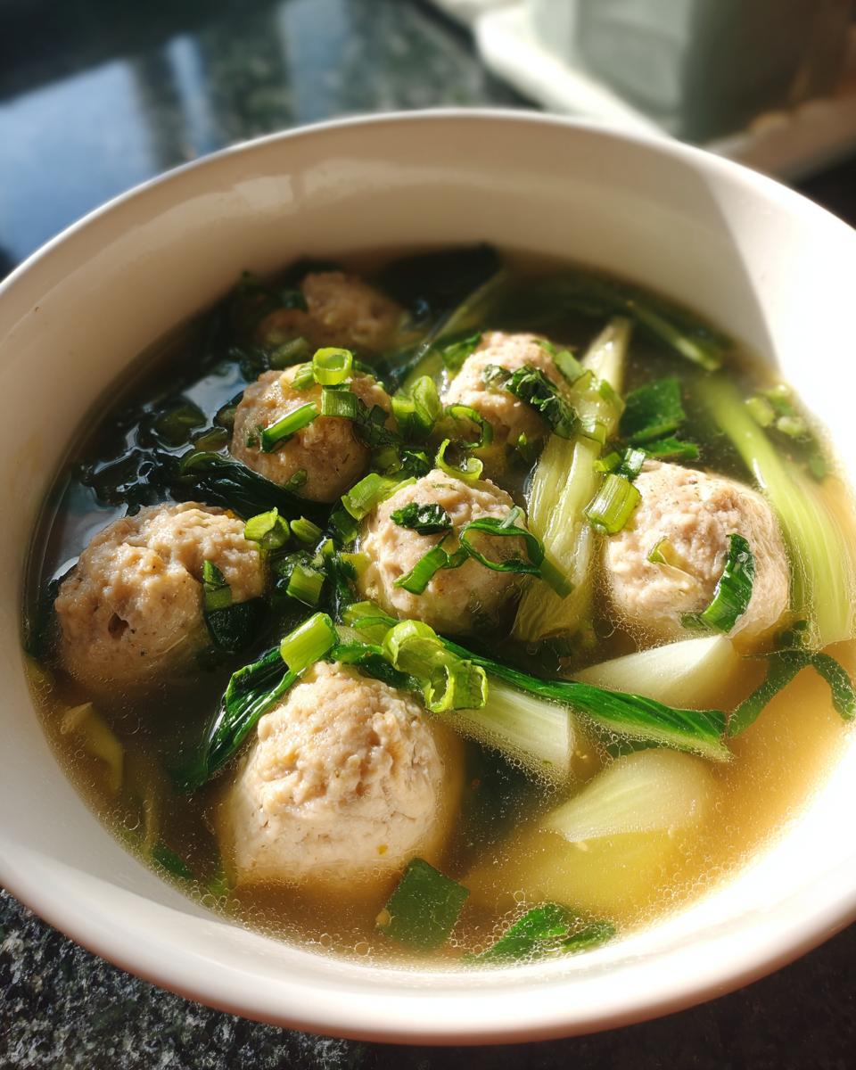 Close-up of a white bowl filled with ginger chicken meatball soup, featuring meatballs, bok choy, and scallions.