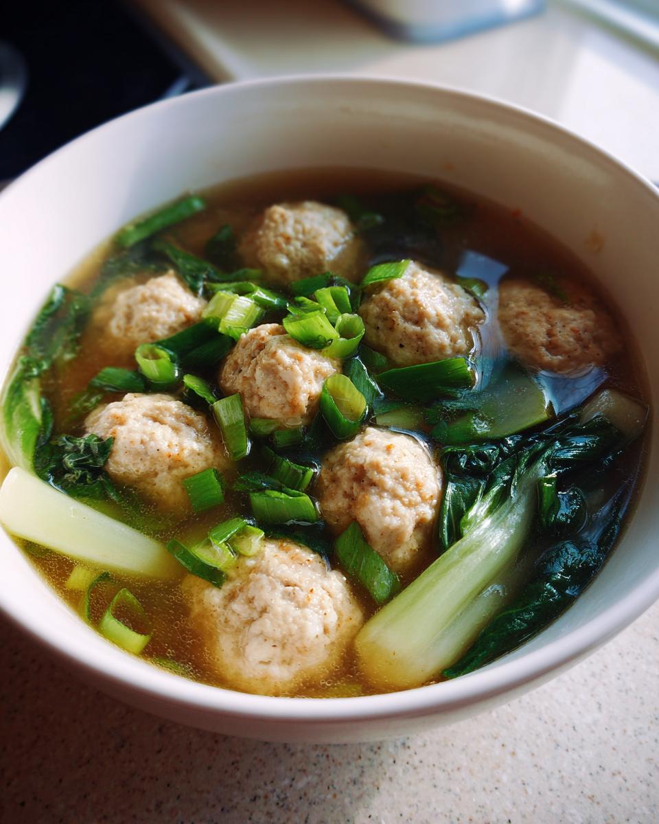 Close-up of a white bowl filled with ginger chicken meatball soup, featuring meatballs, bok choy, and chopped green onions.