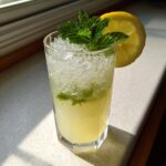 A tall glass filled with a bright yellow ginger lemon cooler, crushed ice, mint leaves, and a lemon slice garnish.