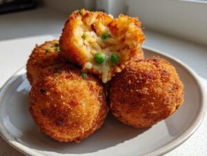 Four golden brown air fryer arancini balls piled on a plate, one is broken open showing rice, melted cheese, and peas.
