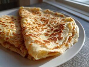 Close-up of folded, golden-brown cottage cheese wrap stacked on a white plate near a window.