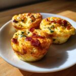 Three golden brown keto egg bites topped with crispy bacon bits and herbs served on a white plate.