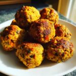 A close-up of several golden-brown vegetarian lentil meatballs piled on a white plate, showing texture and visible vegetable pieces.