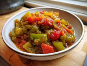 A close-up of chunky, cooked green tomato relish featuring bright green and red pieces in a small ceramic bowl.