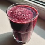 Close-up of a vibrant, deep purple healthy fruit smoothie with kefir, showing foam bubbles on top, sitting near a window.