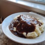 Rich, dark homemade KFC gravy copycat generously poured over fluffy mashed potatoes in a white bowl.