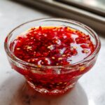 Close-up of bright red, chunky homemade sweet chili sauce with visible chili flakes and sesame seeds in a small glass bowl.
