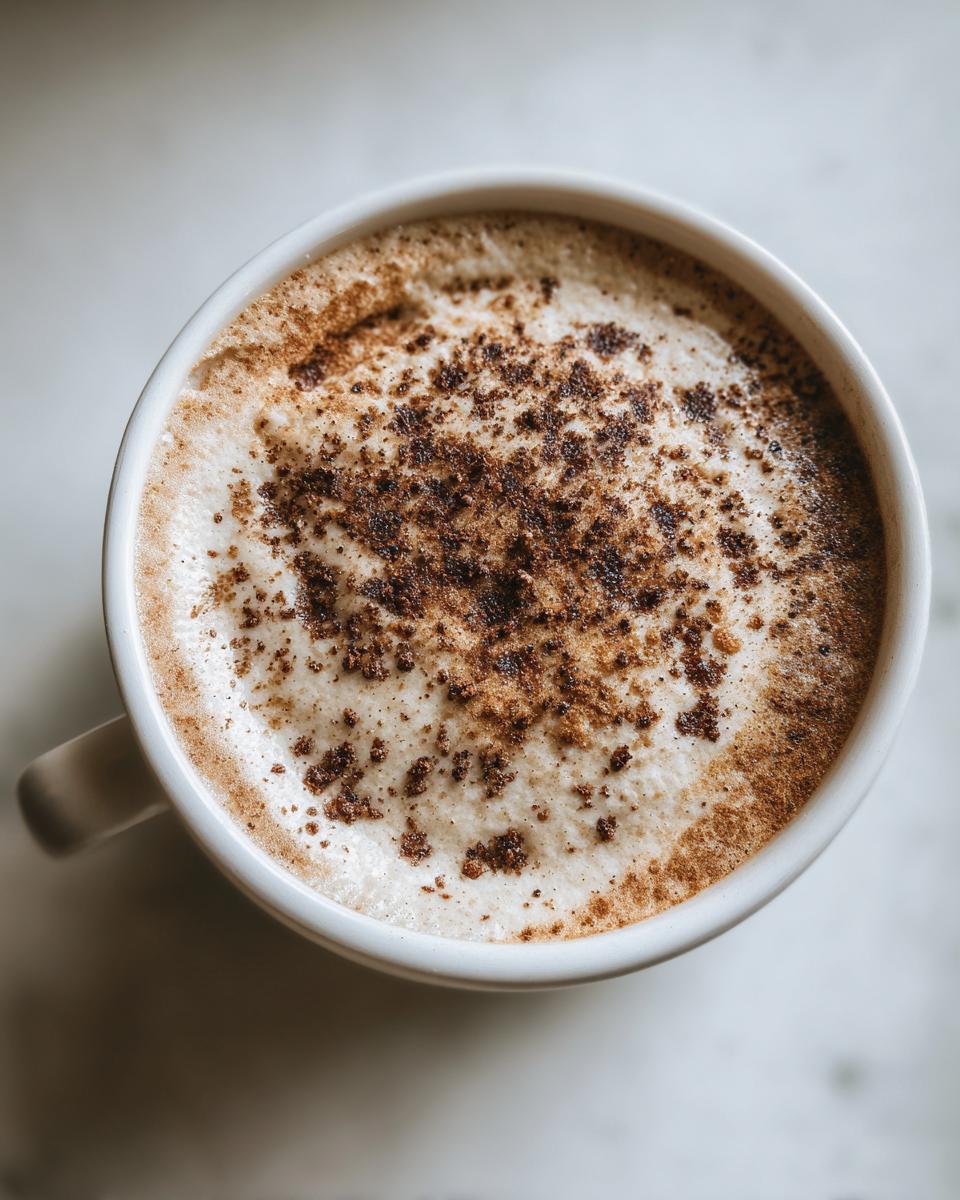 Overhead view of a hot beverage, possibly coffee or tea, topped with foamed milk and sprinkled with dark spices, served in a white mug.