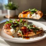 A slice of hot honey ricotta pizza being lifted, with honey dripping over prosciutto, ricotta, and arugula.