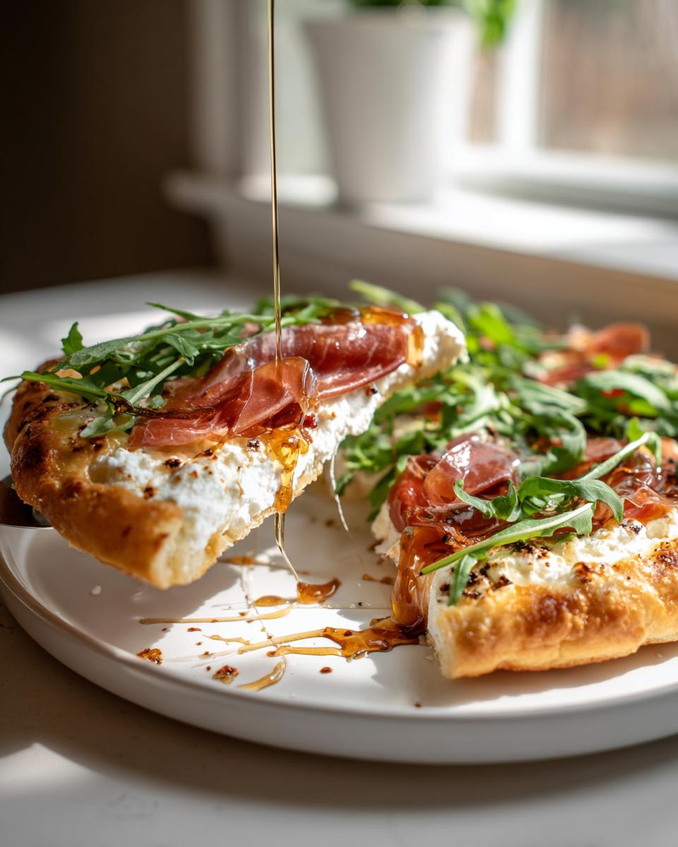 A slice of hot honey ricotta pizza topped with prosciutto and arugula being drizzled with honey.