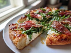 A close-up of a slice of hot honey ricotta pizza topped with prosciutto, arugula, and drizzled with honey.