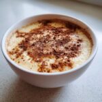 Close-up of creamy jamaican cornmeal porridge with coconut milk topped with ground cinnamon in a white bowl.