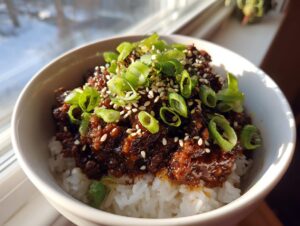 A close-up of a white bowl filled with white rice topped generously with saucy korean ground beef bowl, garnished with sliced green onions and sesame seeds.