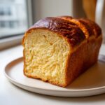 A golden-brown loaf of freshly baked brioche bread, sliced to show its rich, yellow, fluffy interior.