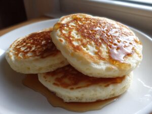 A stack of three fluffy muffin mix pancakes drizzled generously with golden syrup on a white plate.
