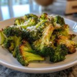 A close-up of perfectly roasted broccoli florets topped with grated Parmesan cheese on a white plate.