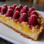 A close-up cross-section of a raspberry mango tart showing the yellow mango filling, crust, and fresh raspberries on top.