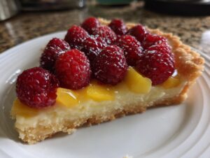 Close-up of a slice of raspberry mango tart showing the flaky crust, creamy filling, mango pieces, and fresh raspberries on top.