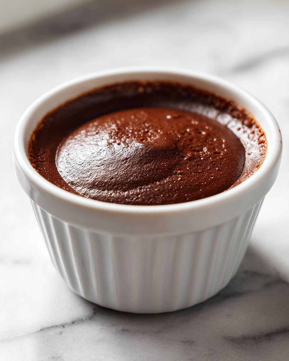 A close-up of rich, dark chocolate custard baked or set in a white, fluted ramekin.