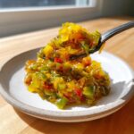 A spoonful of chunky, bright yellow and green tomato relish being lifted from a small white plate.
