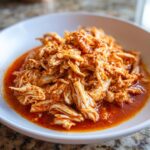 Close-up of shredded chicken tinga coated in a vibrant, rich red chile sauce, served in a white bowl.