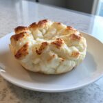 A single, fluffy, baked cloud bread bun with lightly browned peaks sitting on a white plate.