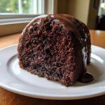 A close-up of a rich, dark slice of dr pepper cake topped with dripping chocolate glaze on a white plate.