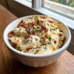 A close-up of a white bowl filled with creamy smoked cream cheese dip, topped generously with crumbled bacon and green onions.