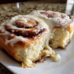 A close-up of a soft, freshly baked gluten-free cinnamon roll covered in thick white icing, with a piece pulled away.