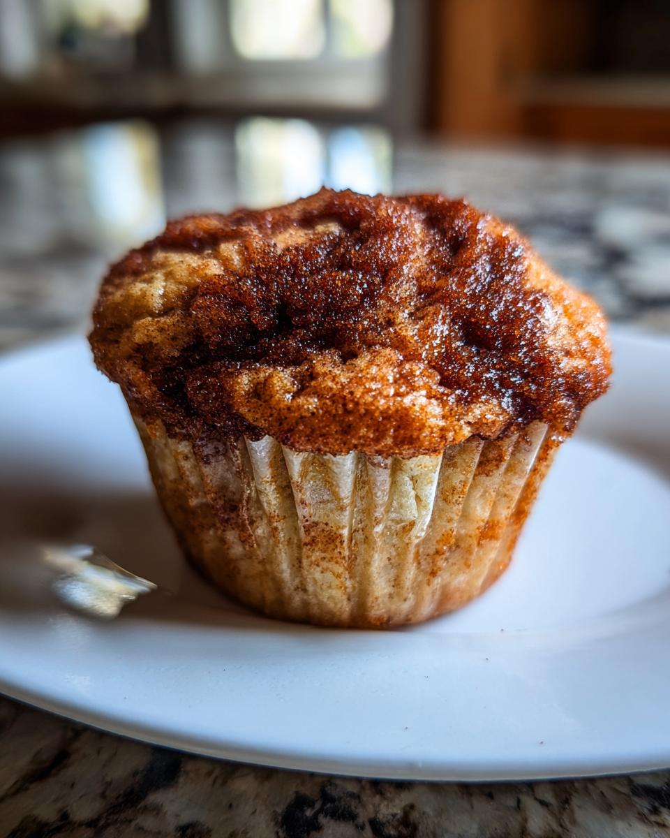 A single, moist sourdough discard cinnamon muffin with a dark, sugary cinnamon topping, sitting on a white plate.